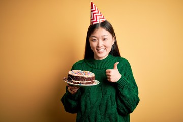 Young asian woman holding birthday cake wearing party hat over yellow isolated background happy with big smile doing ok sign, thumb up with fingers, excellent sign