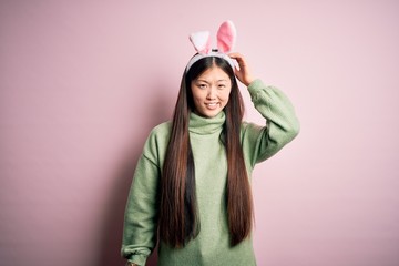 Wall Mural - Young asian woman wearing cute easter bunny ears over pink background confuse and wonder about question. Uncertain with doubt, thinking with hand on head. Pensive concept.