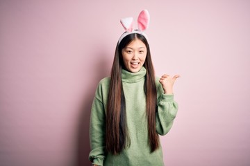 Canvas Print - Young asian woman wearing cute easter bunny ears over pink background smiling with happy face looking and pointing to the side with thumb up.