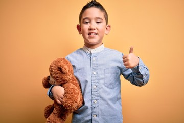 Young little boy kid hugging teddy bear stuffed animal over yellow background happy with big smile doing ok sign, thumb up with fingers, excellent sign