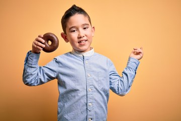 Sticker - Young little boy kid eating unhealthy chocolate doughnut over isolated yellow background very happy pointing with hand and finger to the side