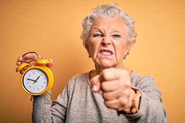 Senior beautiful woman holding alarm clock standing over isolated yellow background annoyed and frustrated shouting with anger, crazy and yelling with raised hand, anger concept