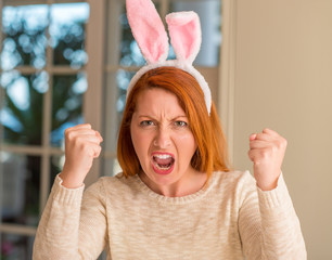 Poster - Redhead woman wearing easter rabbit ears at home annoyed and frustrated shouting with anger, crazy and yelling with raised hand, anger concept