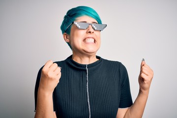 Sticker - Young woman with blue fashion hair wearing thug life sunglasses over white background very happy and excited doing winner gesture with arms raised, smiling and screaming for success. Celebration