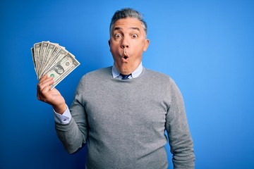 Poster - Middle age handsome grey-haired man holding bunch of dollars over blue background scared in shock with a surprise face, afraid and excited with fear expression