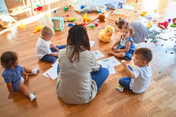 Beautiful teacher and group of toddlers sitting on the floor drawing using paper and pencil around lots of toys at kindergarten