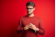 © Krakenimages.com - Young handsome man with beard wearing glasses and sweater standing over red background Hands together and fingers crossed smiling relaxed and cheerful. Success and optimistic