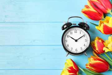 Poster - Black alarm clock with spring flowers and space for text on light blue wooden background, flat lay. Time change