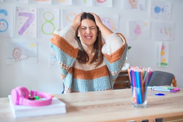 Sticker - Young beautiful teacher woman wearing sweater and glasses sitting on desk at kindergarten suffering from headache desperate and stressed because pain and migraine. Hands on head.