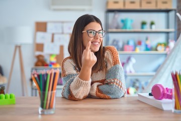 Poster - Young beautiful teacher woman wearing sweater and glasses sitting on desk at kindergarten Beckoning come here gesture with hand inviting welcoming happy and smiling