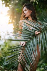 Wall Mural - slim slender lady covering perfect body with green plant, female with dark hair look side, stand in tropical rainforest. green swimsuit is behind green leaf. wildlife, forest, people concept