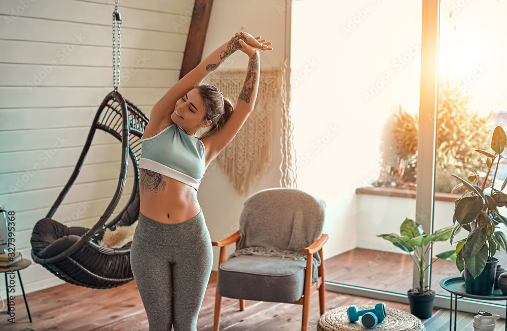Fitness woman exercising and stretching arms at home in the living room in sportswear. Female doing relaxing yoga and breathing exercises. - obrazy, fototapety, plakaty 