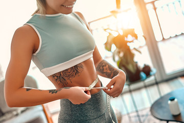 Weight loss, slim body, healthy lifestyle concept. Cropped image of a slim young woman measuring her thin waist with a tape measure at home in the living room, close up.