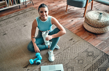 A sporty woman in sportswear is sitting on the floor with dumbbells and a protein shake or a bottle of water and is using a laptop at home in the living room. Sport and recreation concept.