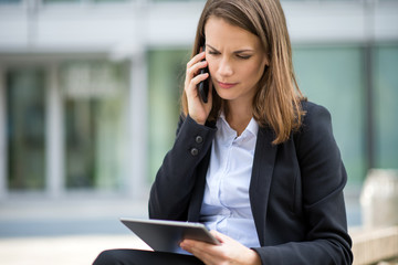 Woman using a tablet while talking on her mobile cell phone in front of her office