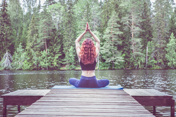 Wall Mural - Young yogi  girl  practicing yoga, sitting in variation of Gomukhasana, Cow Face Posture  on the pier of a beautiful lake. Concept of healthy life and natural balance