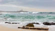 © wewi-creative - A tree trunk lies on the shore in the north of Spain in Galicia on the sand in front of a stone. In the background the Larino lighthouse on the Atlantic coast. Big waves on the beach.