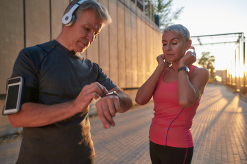 Wall Mural - Best music for training. Healthy and fit middle-aged couple in sport clothing adjusting headphones and setting their playlist while exercising together outdoors