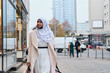 Young stylish Arabic woman in hijab dreamily walking around street with shopping bags