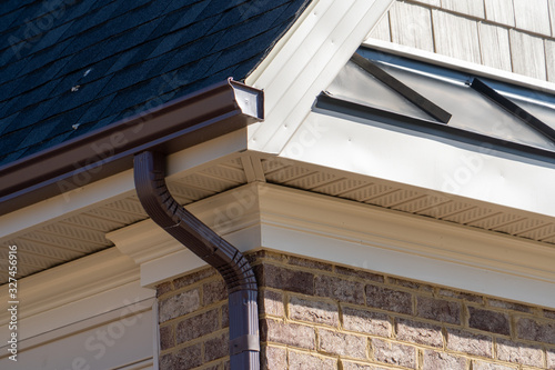 Closeup view of dark brown gutter system with white soffit vent, gutter guard, drop outlet, downspout, vinyl elbows, decorative trim molding, on the corner of a brick luxury house in America
