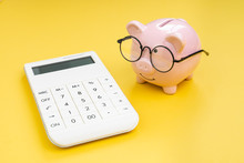 Piggy Bank Wearing Glasses Looking At Calculator On Yellow Background Using As Budget, Tax Cost And Expense Calculation Or Money And Finance Concept