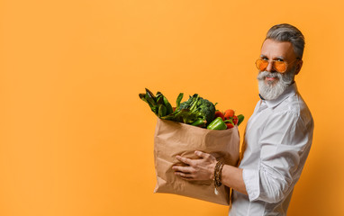 Cool old mature senior man with gray beard shopping hold grocery shopping bag with healthy organic vegetables on yellow