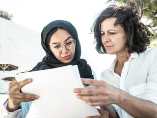 Puzzled female colleagues checking project papers. Multicultural businesswomen in office clothing and Arabian veil reading documents outside. Paperwork error concept