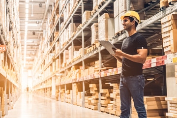 Smart Indian engineer man worker wearing safety helmet doing stocktaking of product management in cardboard box on shelves in warehouse. Factory physical inventory count.