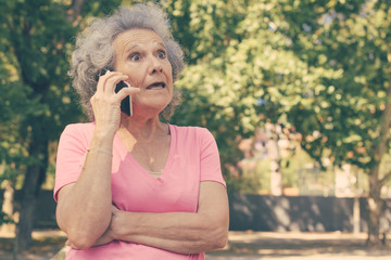 Excited surprised old lady getting shocking news from phone call. Senior grey haired woman with wide eyes standing outdoors and talking on mobile phone. Shock concept