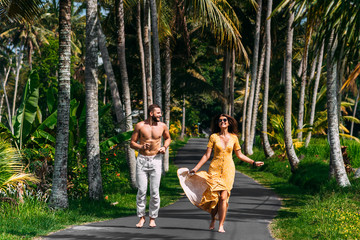 Beautiful couple running among palm trees in Bali, Indonesia. Honeymoon on the Islands. A happy couple in love runs along the road. Honeymoon trip. Wedding trip. Couple on vacation in Bali