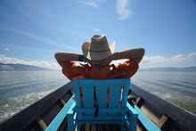 Young Man In Hat Sitting On Chair In Boat With Hands Behind Head And Enjoying Life, Blue Sky And Clean Water Of Lake