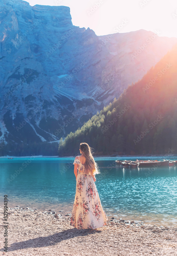 Girl with hair flying in wind red-haired, long silk elegancy dress floral print turned away stands on shore Lake Bryce enjoy silence beauty sunshine mountains. Backdrop boat water mountain with forest - obrazy, fototapety, plakaty 
