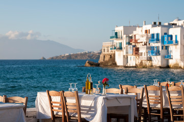 Wall Mural - Bright scenic afternoon view of empty tables waiting for sunset diners set up along the harbor in the old town of Mykonos, Greece