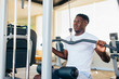 © twinsterphoto - Strong African American man in sportswear pulling cable with bar while sitting on exercise machine during training in gym.