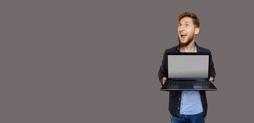 Ginger caucasian man with beard is looking up to gray blank space while holding a laptop and advertising something