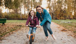 © Strelciuc - Caucasian mother looking through her eyeglasses is helping her daughter to ride the bike during an autumn walk