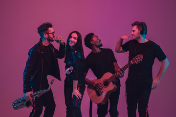 Group of young multiethnic musicians created band, dancing in neon light on pink background. Concept of music, hobby, festival, wellness. Joyful party host, dancer, singer, guitarist, saxophonist.