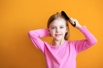 Cute baby, baby girl 6-8 years old combs her soft blond hair medium length comb and smiles on an orange background