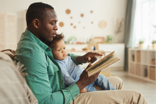 Side View Portrait Of Caring African-American Dad Reading Book To Cute Baby Son At Home, Copy Space