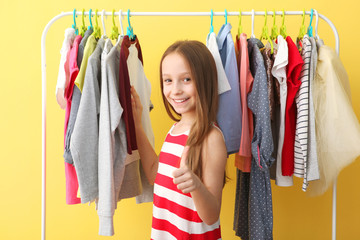 Wall Mural - Cute cheerful little girl chooses clothes with floor hangers. Children's clothing, children's shopping. Fashionable clothes for children.