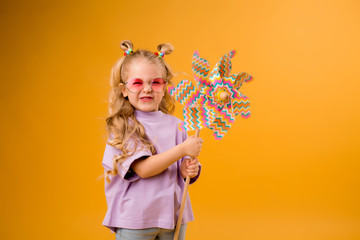 Wall Mural - little blonde girl smiling in sunglasses holding a windmill stands on a yellow background in the Studio isolate. happy child, text space, horizontal photo