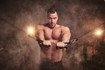 Wall Mural - Muscular and fit male doing an excersise for his pectoral muscles on a hand pull machine in a dark gym surrounded by smoke.