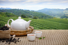 Warm Cup Of Tea With Teapot, Green Tea Leaves And Dried Herbs On The Bamboo Mat At Morning In Plantations Background With Empty Space, Organic Product From The Nature For Healthy With Traditional