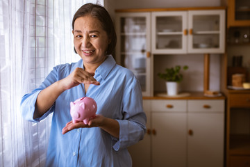 Wall Mural - Asain Elder  putting coin money to piggy bank saving, family, savings, age and people concept - smiling senior couple with money and piggy bank at home.