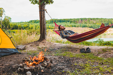 Wall Mural - man laying on hammock at lake beach near camp fire