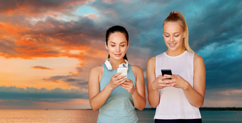 Wall Mural - fitness, sport and healthy lifestyle concept - smiling young women or female friends with smartphones over sea and sunset sky on background