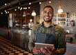 © StratfordProductions - Young african-american man wearing apron standing using digital tablet in coffee shop