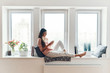 Carefree young woman in cozy pajamas using smart phone and smiling while resting on the window sill at home