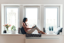 Carefree Young Woman In Cozy Pajamas Reading A Book While Resting On The Window Sill At Home