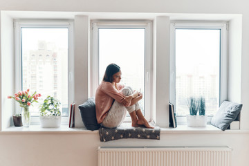 Wall Mural - Attractive young woman in cozy pajamas thinking about something while resting on the window sill at home